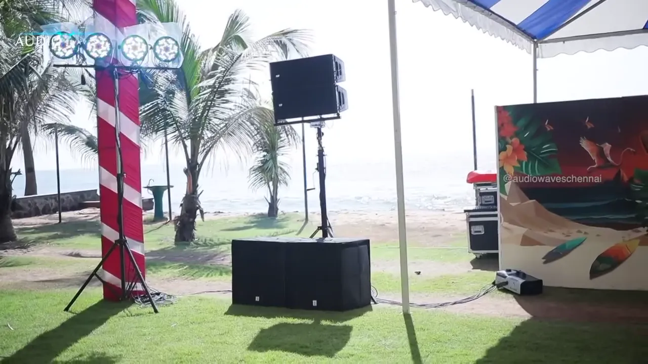 Open-air speakers on stands beside a tropical stage backdrop by the beach
